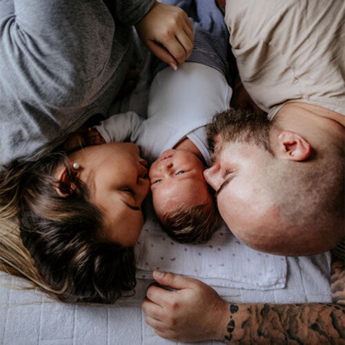 Una mamá, papá y su bebé tumbados sobre la cama besando al bebé que esta entre los dos. - Mustela España - 1