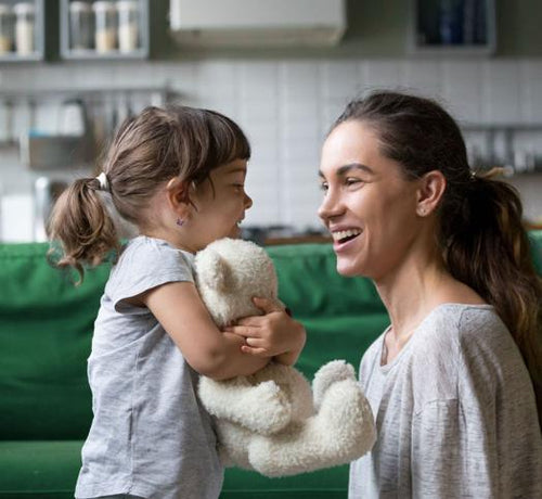 Una niña feliz abraza a su osito mientras le sonríe a su madre en la cocina de su casa - Mustela España - 1