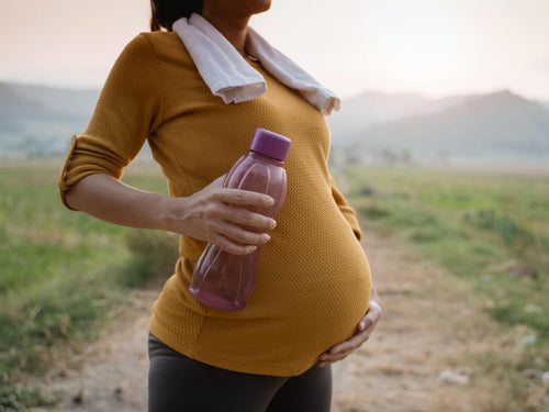 Mujer en la naturaleza con ropa de deporte y botella de agua sujetando su tripa de embarazada  - Mustela España - 1