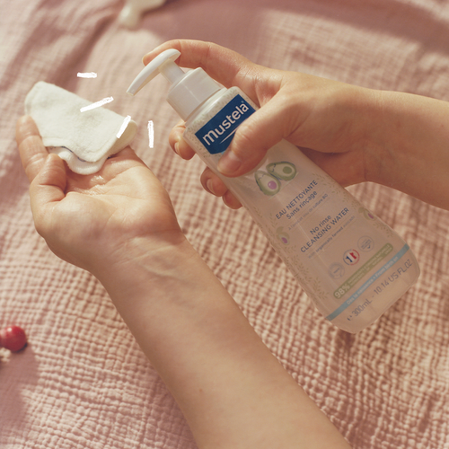 Manos aplicando el agua limpiadora de Mustela sobre un disco de algodón, con una botella con dosificador sobre un fondo rosado. - Mustela España - 1