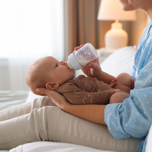 Una madre sentada sobre su cama dando un biberón a su bebé que lo mantiene sostenido sobre sus piernas - Mustela España - 1