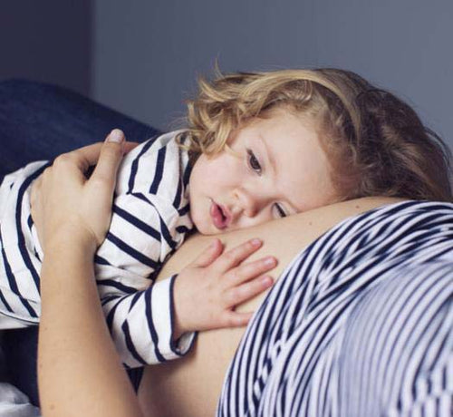 Una bebé abraza el vientre de su madre embarazada mientras las dos están estiradas en la cama - Mustela España - 1